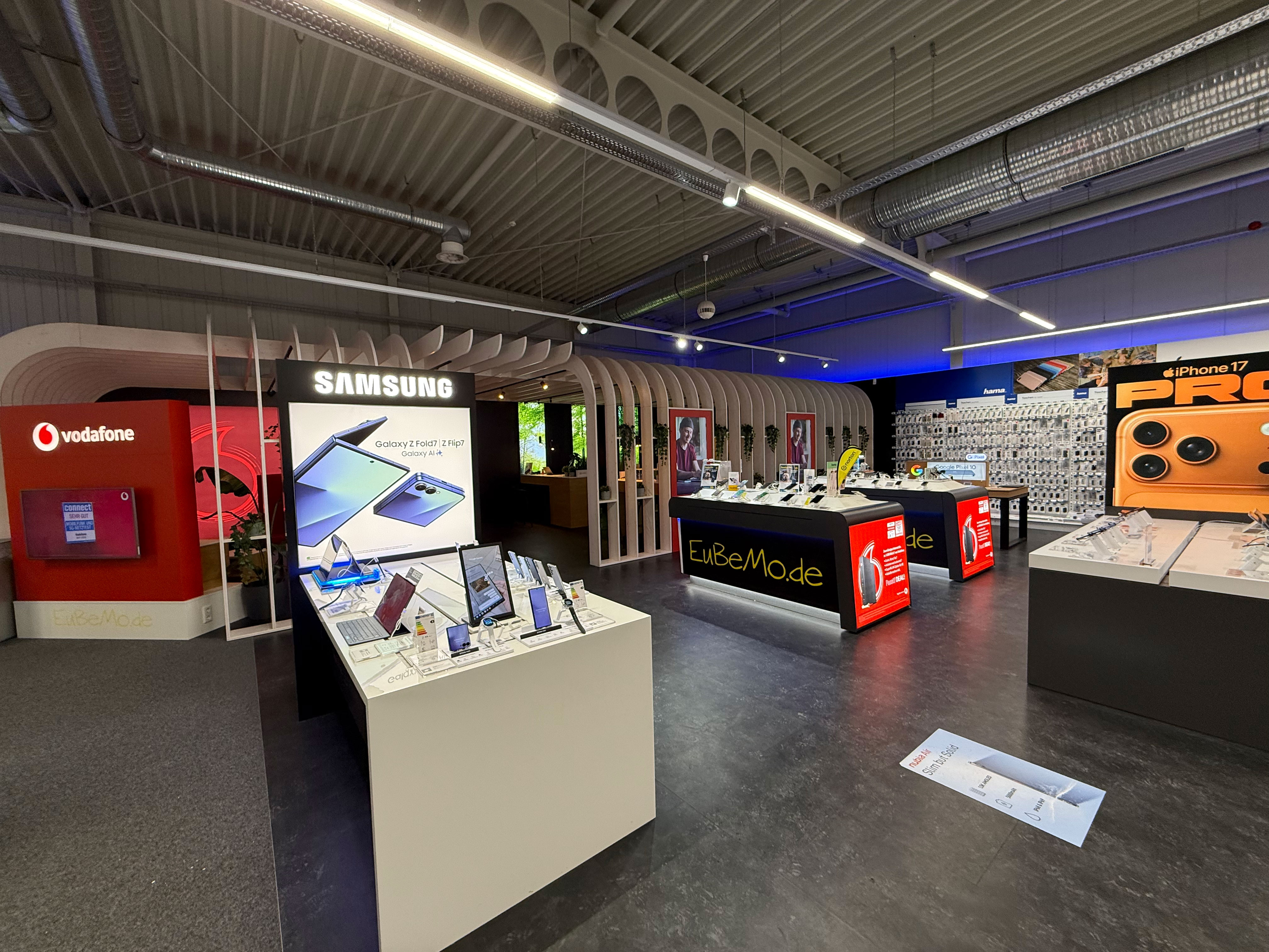Weitwinkelaufnahme einer Mobilfunk-Abteilung im Elektronikmarkt: vorn ein Samsung-Präsentationsstand mit Smartphones und Tablets, links eine rote Vodafone-Fläche, mittig Verkaufstische mit „EuBeMo.de“-Branding. Im Hintergrund eine große Zubehörwand und rechts ein großes Werbebanner „iPhone 17 Pro“ sowie weitere Ausstellungstische.