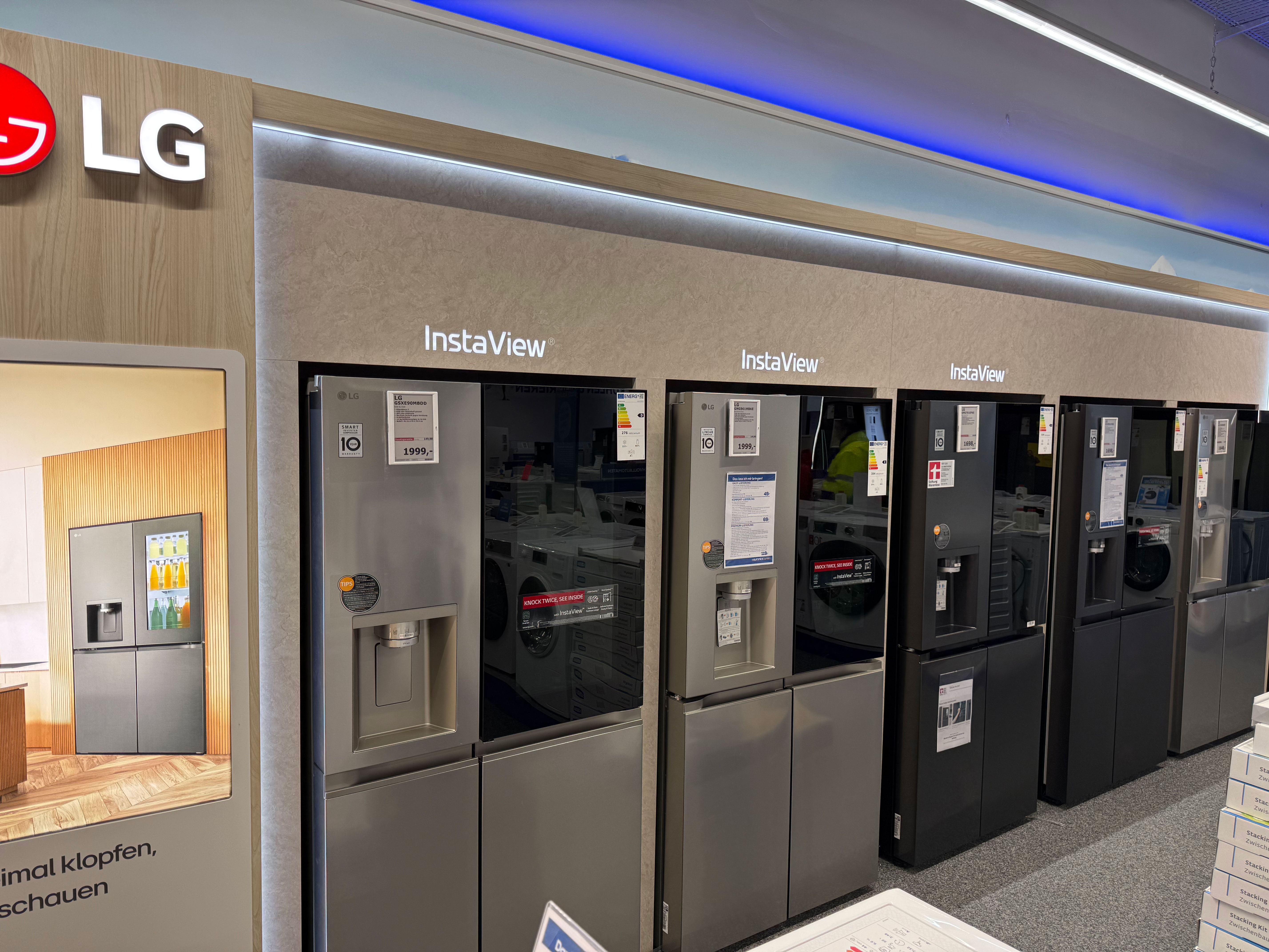 Ausstellungswand mit mehreren LG „InstaView“-Kühlschränken in Grau und Schwarz, teils mit Wasser-/Eisspender und Preisschildern, in einem Elektronikmarkt. Links ist ein LG-Logo und ein Werbedisplay zu sehen, darüber verläuft eine blaue LED-Lichtleiste.