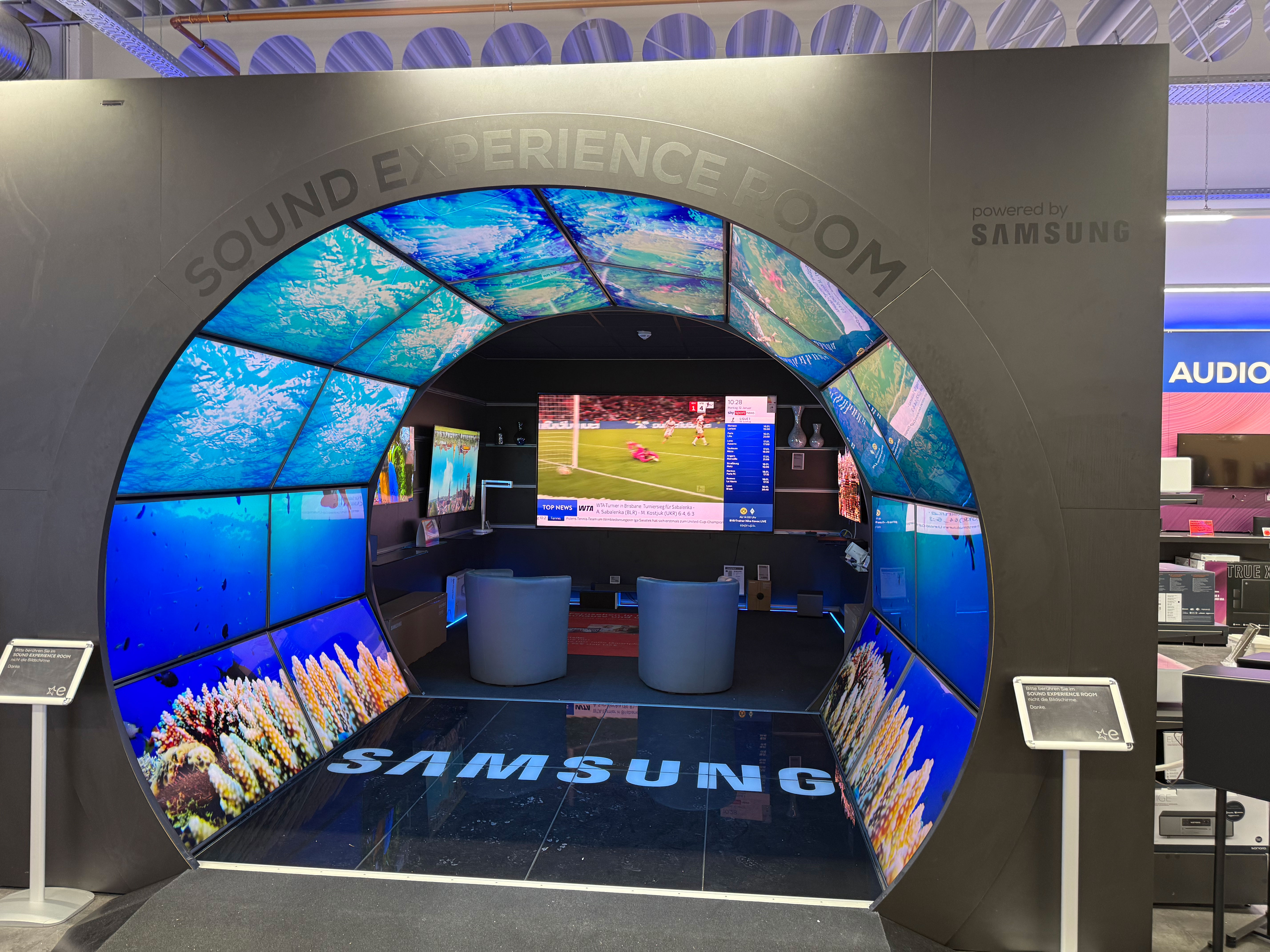 Großer, kreisförmiger „Sound Experience Room“-Eingang mit Samsung-Branding; die Tunnelwände bestehen aus mehreren Displays mit Unterwasserbildern. Im Inneren sind ein großer Fernseher mit Sportübertragung, zwei helle Sessel und Regalflächen zu sehen.