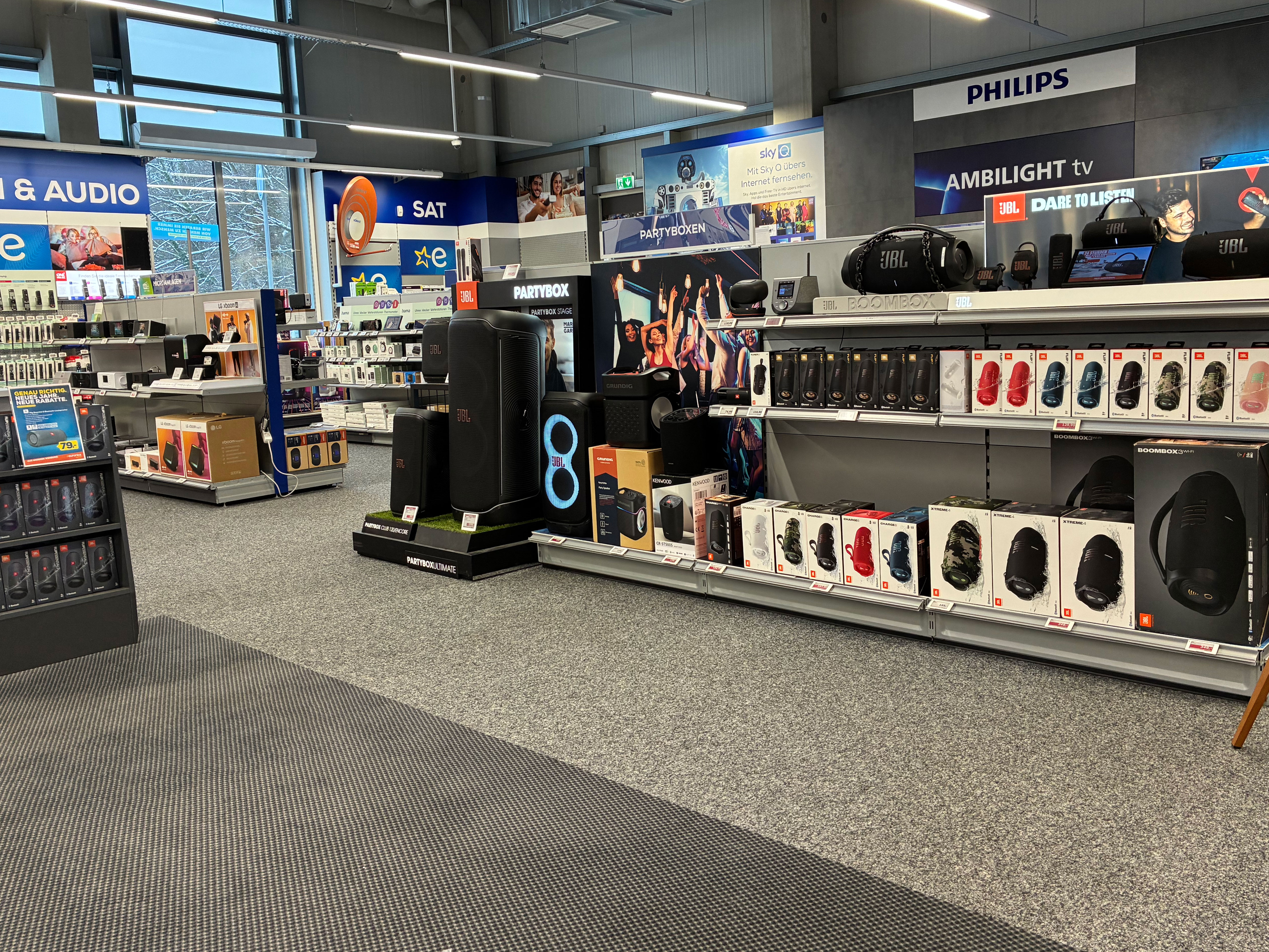 Audio-Abteilung im Elektrofachmarkt: Vorne steht eine JBL-Partybox-Präsentation mit großen Lautsprechern, daneben ein Regal mit zahlreichen JBL-Boxen und Kopfhörern in Verpackungen. Im Hintergrund sind weitere Bereiche wie „SAT“ und „TV & Audio“ sowie ein Philips-Ambilight-TV-Banner zu sehen.