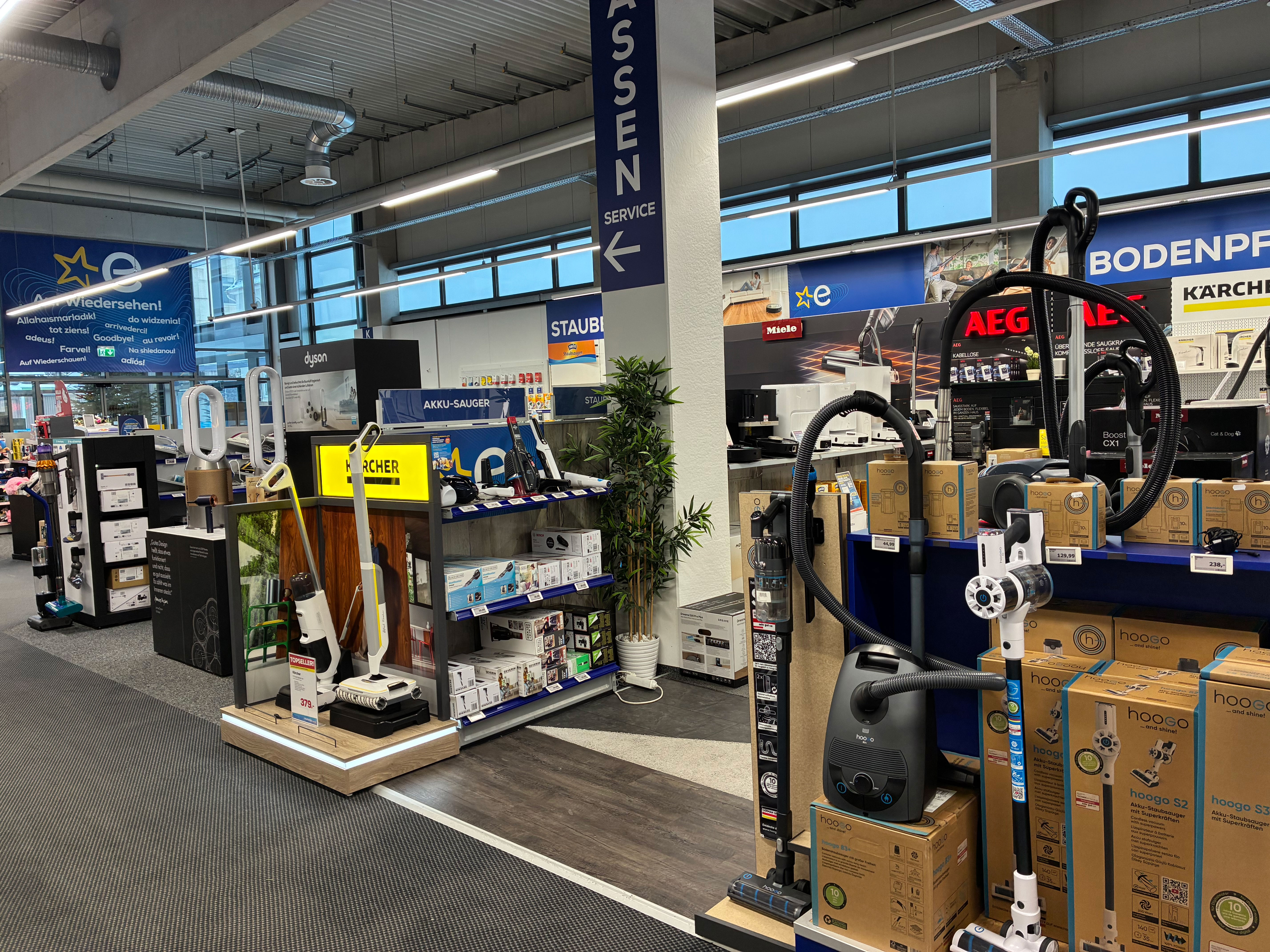 Blick in eine helle Elektrofachmarkt-Abteilung für Staubsauger: Vorne stehen mehrere Akku- und Bodenstaubsauger auf Präsentationsflächen, rechts ein AEG-Display mit verpackten Geräten, links u. a. Dyson und Kärcher; darüber hängen Schilder wie „KASSEN“ und „AKKU-SAUGER“ sowie Markenbanner (z. B. Miele, Kärcher).