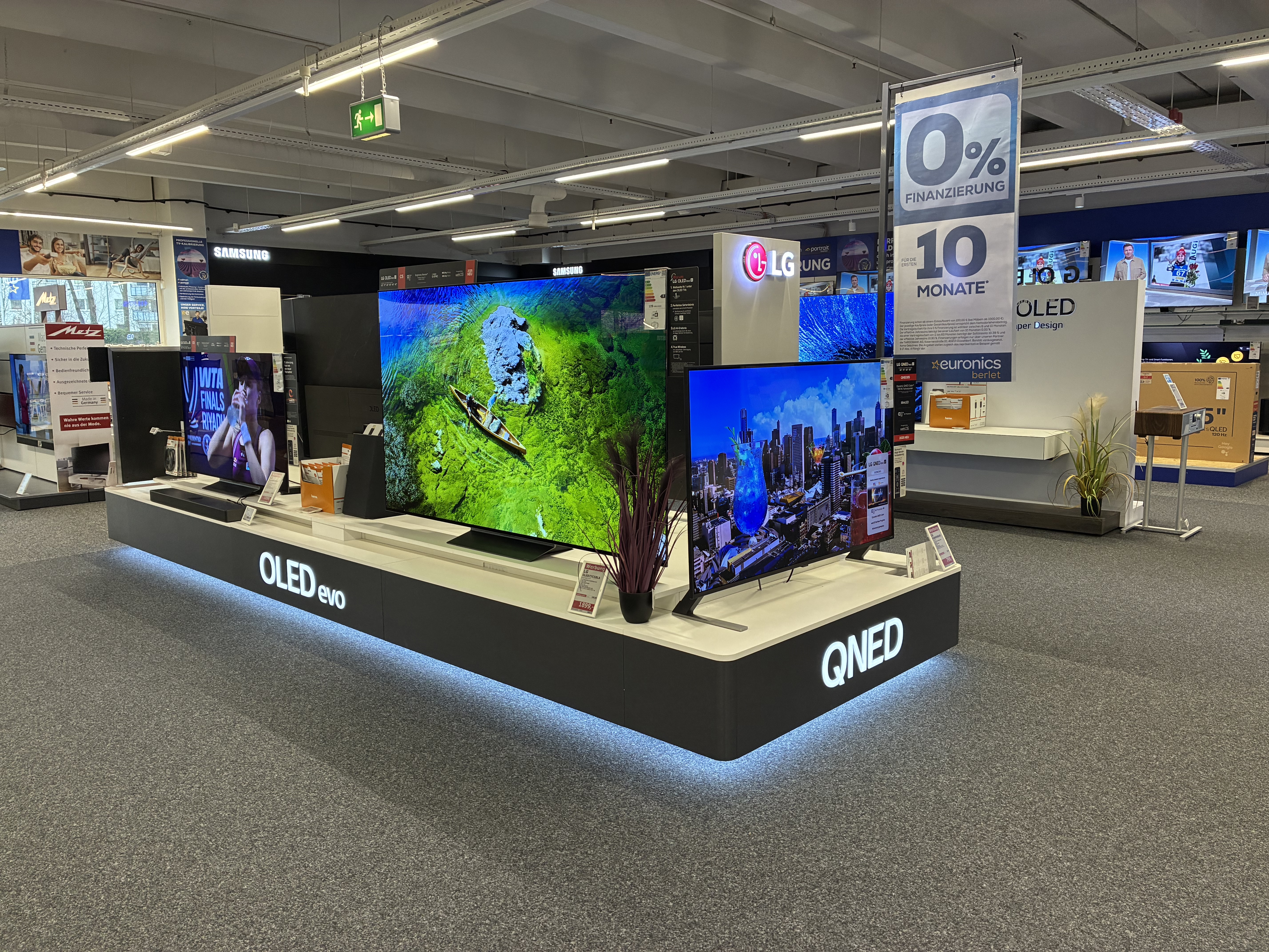 TV-Ausstellungsfläche im Elektrofachmarkt: Auf einem beleuchteten Podest stehen mehrere große Fernseher mit farbigen Demo-Bildern, daneben Markenbeschriftungen wie „OLED evo“ und „QNED“. Im Hintergrund sind weitere TV-Wände (u. a. Samsung/LG) zu sehen sowie ein großes Banner „0% Finanzierung – 10 Monate“.