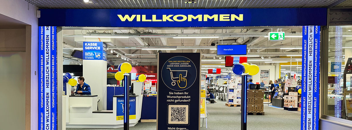 Eingang des EURONICS Berlet Marktes mit großem blauen „Willkommen“-Schriftzug, gelb-blauen Luftballons und Hinweisschild für Online-Bestellungen und Click & Collect Service.