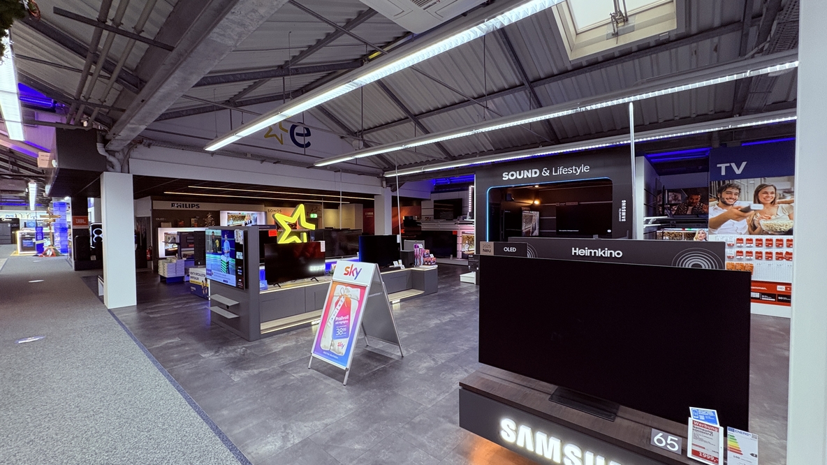 TV- und Heimkinoabteilung bei EURONICS-Berlet-Bergkamen mit großem Samsung-Fernseher im Vordergrund, Sky-Werbeständer in der Mitte und dahinter einem modern beleuchteten „Sound & Lifestyle“-Bereich mit weiteren Fernsehern und Markenflächen. TV- und Heimkinoabteilung bei EURONICS-Berlet-Bergkamen mit großem Samsung-Fernseher im Vordergrund, Sky-Werbeständer in der Mitte und dahinter einem modern beleuchteten „Sound & Lifestyle“-Bereich mit weiteren Fernsehern und Markenflächen.