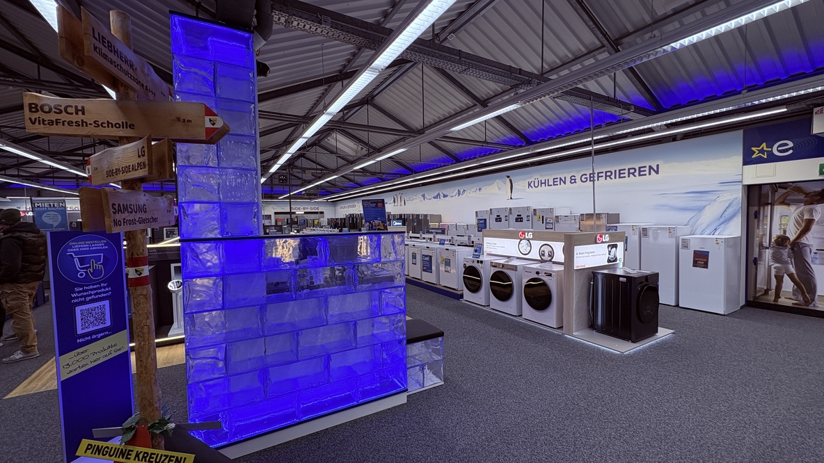 Dekorativ gestaltete Kühl- und Gefriergeräteabteilung bei EURONICS-Berlet-Bergkamen mit leuchtender blauer „Eiswand“, Holzwegweiser zu Marken wie Liebherr, Bosch, LG und Samsung sowie Reihen von Waschmaschinen, Kühlschränken und Gefriertruhen im Hintergrund. Dekorativ gestaltete Kühl- und Gefriergeräteabteilung bei EURONICS-Berlet-Bergkamen mit leuchtender blauer „Eiswand“, Holzwegweiser zu Marken wie Liebherr, Bosch, LG und Samsung sowie Reihen von Waschmaschinen, Kühlschränken und Gefriertruhen im Hintergrund.