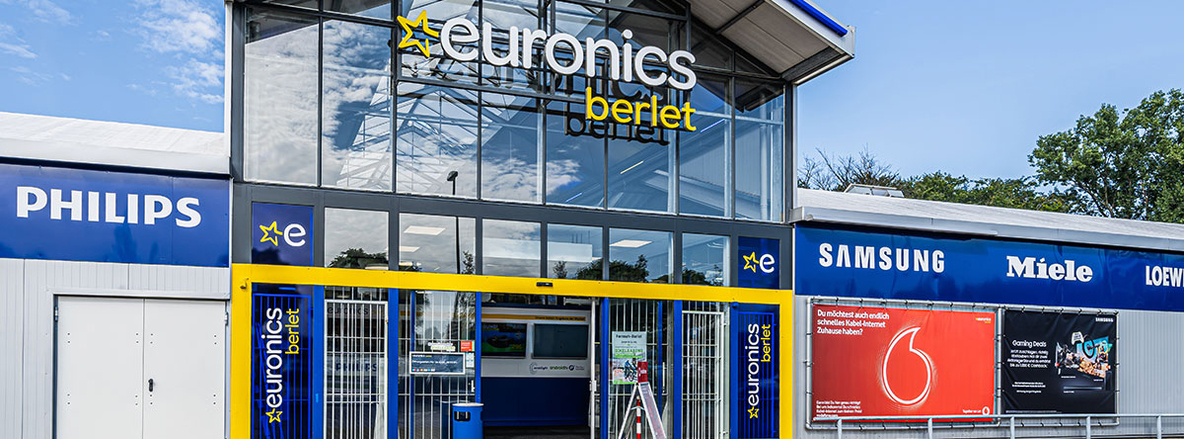 Außenaufnahme der Euronics-Berlet-Filiale in Bergkamen: Ein hoher Glasportikus mit dunklem Stahlrahmen, darüber das Leuchtschild „euronics berlet“ in Weiß und Gelb. Rechts und links schieben sich blaue Fassadenbänder mit Markenlogos wie Philips, Samsung und Miele vorbei. Der automatische Doppeltür-Eingang ist von einer gelben Rahmung umgeben, im Vordergrund ein Poller und ein Vodafone-Werbebanner neben dem rechten Flügel. Im Spiegelglas zeichnen sich blauer Himmel und Baumkronen ab. Außenaufnahme der Euronics-Berlet-Filiale in Bergkamen: Ein hoher Glasportikus mit dunklem Stahlrahmen, darüber das Leuchtschild „euronics berlet“ in Weiß und Gelb. Rechts und links schieben sich blaue Fassadenbänder mit Markenlogos wie Philips, Samsung und Miele vorbei. Der automatische Doppeltür-Eingang ist von einer gelben Rahmung umgeben, im Vordergrund ein Poller und ein Vodafone-Werbebanner neben dem rechten Flügel. Im Spiegelglas zeichnen sich blauer Himmel und Baumkronen ab.