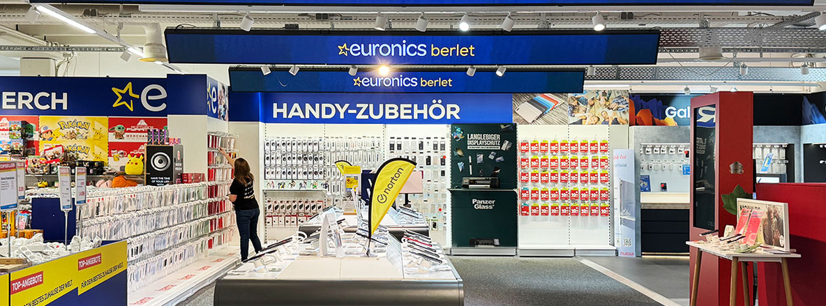 Handy-Zubehör-Abteilung im EURONICS Berlet Markt mit Schildern, Smartphone-Hüllen, Panzerfolien und Zubehörartikeln; im Vordergrund gelbe ynoton-Flagge. Handy-Zubehör-Abteilung im EURONICS Berlet Markt mit Schildern, Smartphone-Hüllen, Panzerfolien und Zubehörartikeln; im Vordergrund gelbe ynoton-Flagge.