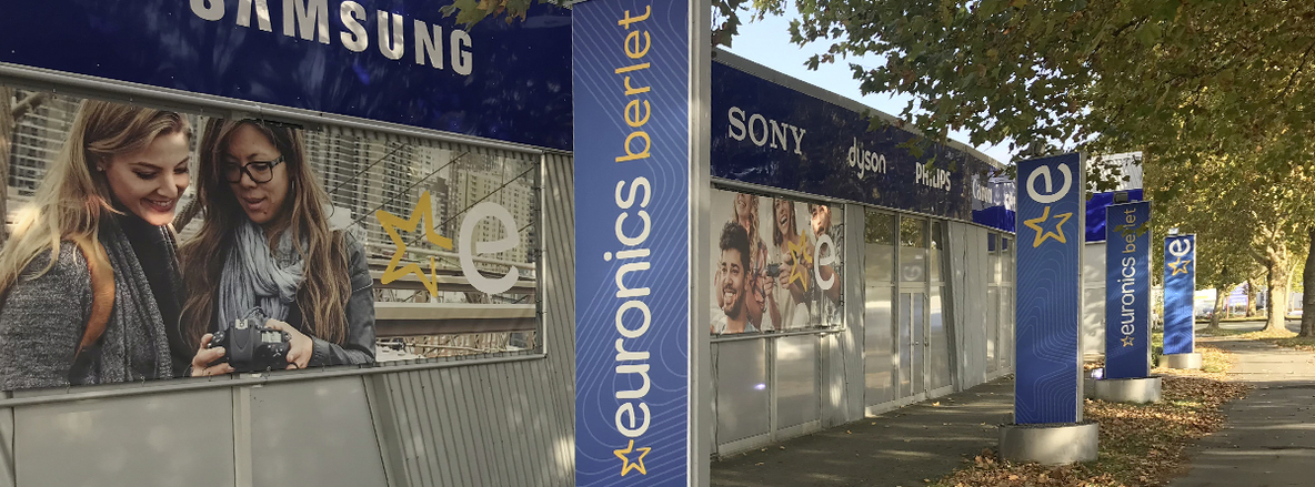 Außenansicht einer Euronics-Berlet-Filiale entlang eines von Laubbäumen gesäumten Gehwegs: Rechts reihen sich mehrere hohe Stehlen in Blau mit dem weißen „euronics berlet“-Schriftzug und gelbem Sternsymbol; sie stehen auf dem Gehsteig. Dahinter verläuft eine lange Gebäudefassade mit silbergrauen Metallpaneelen und großflächigen Fensterfronten, darüber ein dunkelblaues Fassadenband mit leuchtenden Markenlogos („SONY“, „dyson“, „PHILIPS“, „Canon“). Auf den Glasflächen sind großflächige Werbefolien mit jungen Menschen drauf zu sehen. Außenansicht einer Euronics-Berlet-Filiale entlang eines von Laubbäumen gesäumten Gehwegs: Rechts reihen sich mehrere hohe Stehlen in Blau mit dem weißen „euronics berlet“-Schriftzug und gelbem Sternsymbol; sie stehen auf dem Gehsteig. Dahinter verläuft eine lange Gebäudefassade mit silbergrauen Metallpaneelen und großflächigen Fensterfronten, darüber ein dunkelblaues Fassadenband mit leuchtenden Markenlogos („SONY“, „dyson“, „PHILIPS“, „Canon“). Auf den Glasflächen sind großflächige Werbefolien mit jungen Menschen drauf zu sehen.