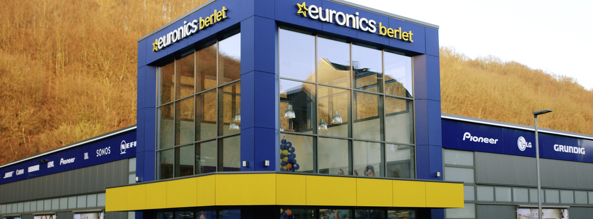 Außenansicht von Euronics-Berlet-Ennepetal: Ein zweigeschossiger Kubus mit markanten, blauen Eckpfeilern und großflächigen Glasfenstern im Obergeschoss, darüber das leuchtende „euronics berlet“-Logo. An der umlaufenden Fassadenblende in Dunkelblau sind Logos bekannter Marken wie Pioneer, LG und Grundig zu sehen. Im Hintergrund ein sanft bewachsener Hügel, im Vordergrund eine gelbe Fassadenblende über dem Eingang. Außenansicht von Euronics-Berlet-Ennepetal: Ein zweigeschossiger Kubus mit markanten, blauen Eckpfeilern und großflächigen Glasfenstern im Obergeschoss, darüber das leuchtende „euronics berlet“-Logo. An der umlaufenden Fassadenblende in Dunkelblau sind Logos bekannter Marken wie Pioneer, LG und Grundig zu sehen. Im Hintergrund ein sanft bewachsener Hügel, im Vordergrund eine gelbe Fassadenblende über dem Eingang.