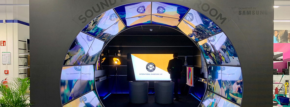 Begehbare „Sound Room“-Installation bei Euronics Berlet in Arnsberg-Neheim: Ein großer, schwarzer halbrunder Torbogen, belegt mit rechteckigen, gebogenen Displays, die bunte Videoinhalte zeigen. Durch den Bogen blickt man auf zwei graue Sessel vor einem großen Bildschirm mit dem Schriftzug „International Champions Cup“. Ein Mann in Anzug steht seitlich vor dem Screen, im Hintergrund Regale mit Elektrogeräten und weiße Deckenrasterbeleuchtung. Begehbare „Sound Room“-Installation bei Euronics Berlet in Arnsberg-Neheim: Ein großer, schwarzer halbrunder Torbogen, belegt mit rechteckigen, gebogenen Displays, die bunte Videoinhalte zeigen. Durch den Bogen blickt man auf zwei graue Sessel vor einem großen Bildschirm mit dem Schriftzug „International Champions Cup“. Ein Mann in Anzug steht seitlich vor dem Screen, im Hintergrund Regale mit Elektrogeräten und weiße Deckenrasterbeleuchtung.