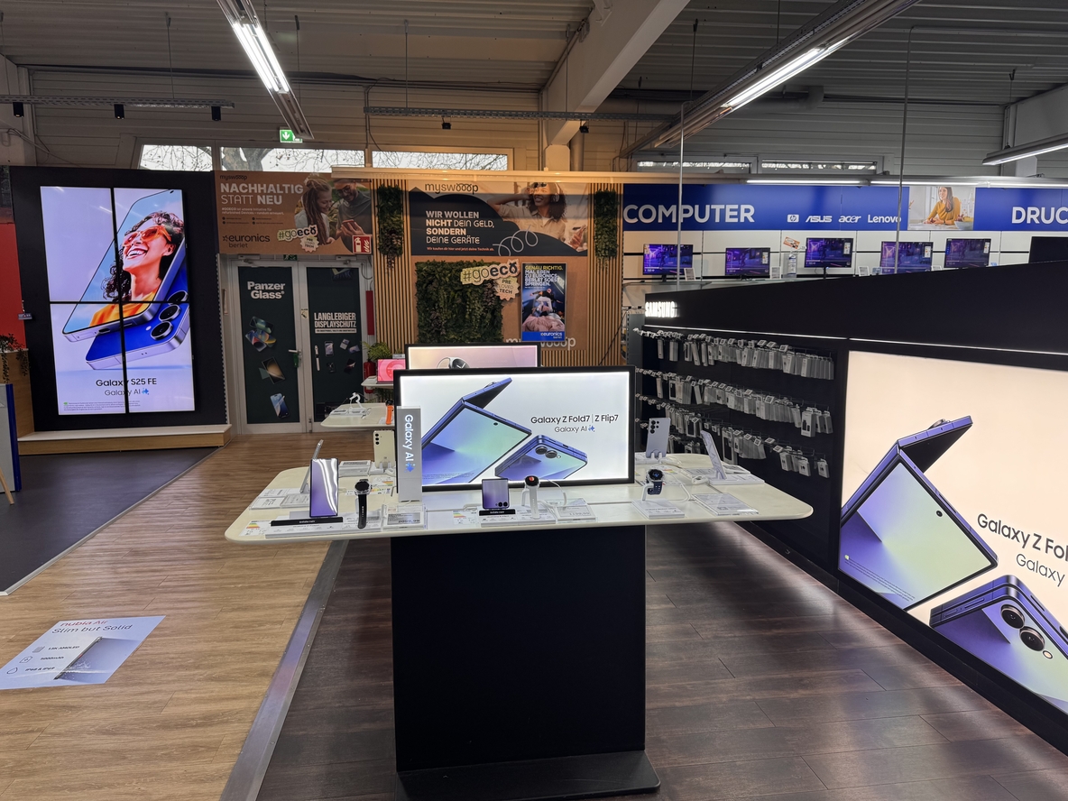 Smartphone-Bereich bei EURONICS-Berlet-Soest mit zentralem Samsung-Galaxy-Präsentationstisch, großen Leuchtdisplays zu Galaxy Z Fold/Z Flip und S25 FE, Zubehörwand rechts sowie Nachhaltigkeits- und mySWOOP-Werbeflächen im Hintergrund. Smartphone-Bereich bei EURONICS-Berlet-Soest mit zentralem Samsung-Galaxy-Präsentationstisch, großen Leuchtdisplays zu Galaxy Z Fold/Z Flip und S25 FE, Zubehörwand rechts sowie Nachhaltigkeits- und mySWOOP-Werbeflächen im Hintergrund.