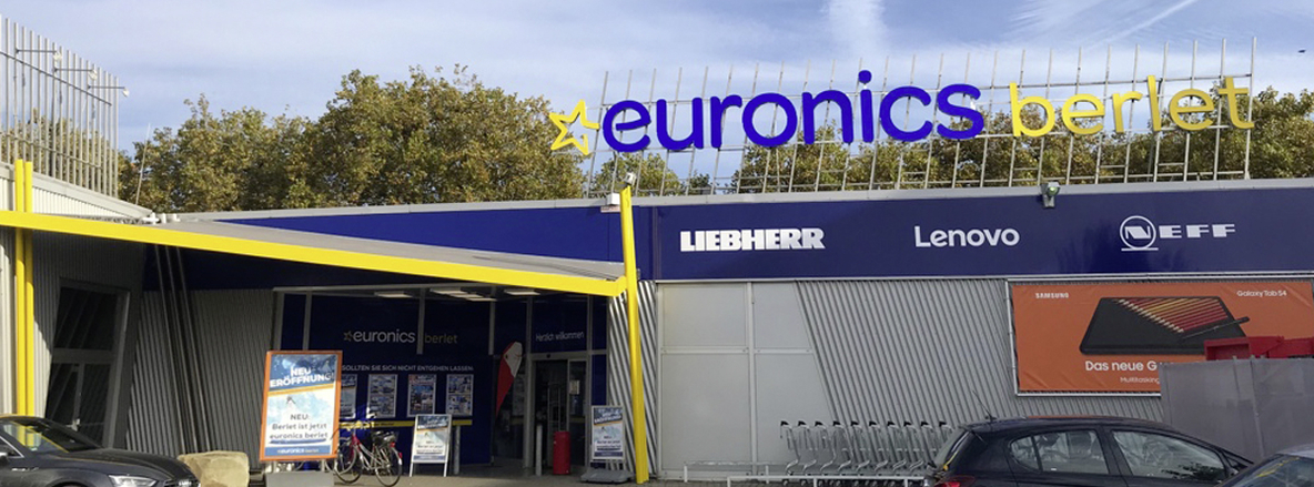 Außenansicht Euronics Berlet Soest mit markanter, schräg nach vorn gezogener gelber Metallüberdachung, die auf den automatischen Doppelflügeltür-Eingang zusteuert. Der obere Fassadenbereich wird von einem großen, leuchtenden „euronics berlet“-Schriftzug auf einem Metallgitter getragen. Darunter verläuft eine blaue Blende mit den Logos von Marken wie Liebherr, Lenovo und NEFF, rechts ist ein orangefarbener Samsung-Werbebanner angebracht. Vor dem Eingangsbereich stehen geordnete Einkaufswagen, im Hintergrund zeichnen sich Laubbäume gegen den klaren Himmel ab. Außenansicht Euronics Berlet Soest mit markanter, schräg nach vorn gezogener gelber Metallüberdachung, die auf den automatischen Doppelflügeltür-Eingang zusteuert. Der obere Fassadenbereich wird von einem großen, leuchtenden „euronics berlet“-Schriftzug auf einem Metallgitter getragen. Darunter verläuft eine blaue Blende mit den Logos von Marken wie Liebherr, Lenovo und NEFF, rechts ist ein orangefarbener Samsung-Werbebanner angebracht. Vor dem Eingangsbereich stehen geordnete Einkaufswagen, im Hintergrund zeichnen sich Laubbäume gegen den klaren Himmel ab.