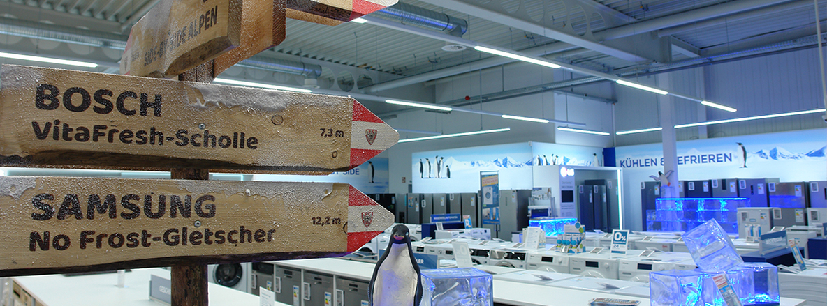 Holzwegweiser-Installation in der Kühlgeräte-Abteilung bei Euronics-Berlet in Arnsberg-Neheim: Zwei grob behauene Schilderpfeile mit roter Spitze, oben „BOSCH VitaFresh-Scholle 7,3 m“, unten „SAMSUNG No Frost-Gletscher 12,2 m“, leicht mit Tau benetzt. Im Hintergrund Reihen weißer Waschmaschinen und silberner Kühlschränke unter weißer Decke, davor ein Deko-Pinguin und blau hinterleuchtete Eiswürfel. Holzwegweiser-Installation in der Kühlgeräte-Abteilung bei Euronics-Berlet in Arnsberg-Neheim: Zwei grob behauene Schilderpfeile mit roter Spitze, oben „BOSCH VitaFresh-Scholle 7,3 m“, unten „SAMSUNG No Frost-Gletscher 12,2 m“, leicht mit Tau benetzt. Im Hintergrund Reihen weißer Waschmaschinen und silberner Kühlschränke unter weißer Decke, davor ein Deko-Pinguin und blau hinterleuchtete Eiswürfel.