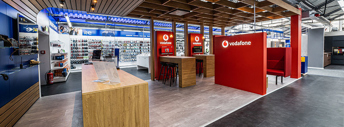 Offener Beratungsbereich mit Vodafone-Lounge bei Euronics-Berlet in Bergkamen: Rechts raumhohe, rote Trennwände mit weißem „vodafone“-Schriftzug, davor zwei erhöhte Holztische mit schwarzen Barhockern, davor eine rote Sitznische mit Polsterbank. Links ein heller Holztisch ohne Stühle, im Hintergrund weiße Regale voller Smartphone-Zubehör unter weißem Deckenraster und blauem LED-Band. Der Boden wechselt von hellem Vinyl im Lounge-Bereich zu dunkelgrauem Vinyl im Markt. Offener Beratungsbereich mit Vodafone-Lounge bei Euronics-Berlet in Bergkamen: Rechts raumhohe, rote Trennwände mit weißem „vodafone“-Schriftzug, davor zwei erhöhte Holztische mit schwarzen Barhockern, davor eine rote Sitznische mit Polsterbank. Links ein heller Holztisch ohne Stühle, im Hintergrund weiße Regale voller Smartphone-Zubehör unter weißem Deckenraster und blauem LED-Band. Der Boden wechselt von hellem Vinyl im Lounge-Bereich zu dunkelgrauem Vinyl im Markt.