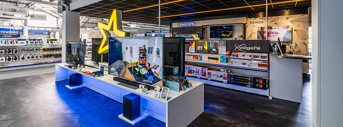 Innenansicht Euronics-Berlet-Bergkamen mit TV- und Audio-Demobereich: Im Vordergrund eine weiße Präsentationstheke mit blauem Unterlicht, darauf ein großer Flachbildfernseher mit Soundbar und Subwoofer. Dahinter steht ein übermannsgroßes, gelb hinterleuchtetes Euronics-Sternlogo. Im rechten Hintergrund ein Philips- und Sonos-Regal mit TV-Zubehör und Vogel’s-Wandhaltern, links eine Wand mit Kopfhörer-Displays. Die Szene ist ausgelegt auf dunkelgrauen Vinylboden und ausgeleuchtet von einem schwarzen Raster-Deckensystem mit warmweißen Leuchtstreifen. Innenansicht Euronics-Berlet-Bergkamen mit TV- und Audio-Demobereich: Im Vordergrund eine weiße Präsentationstheke mit blauem Unterlicht, darauf ein großer Flachbildfernseher mit Soundbar und Subwoofer. Dahinter steht ein übermannsgroßes, gelb hinterleuchtetes Euronics-Sternlogo. Im rechten Hintergrund ein Philips- und Sonos-Regal mit TV-Zubehör und Vogel’s-Wandhaltern, links eine Wand mit Kopfhörer-Displays. Die Szene ist ausgelegt auf dunkelgrauen Vinylboden und ausgeleuchtet von einem schwarzen Raster-Deckensystem mit warmweißen Leuchtstreifen.