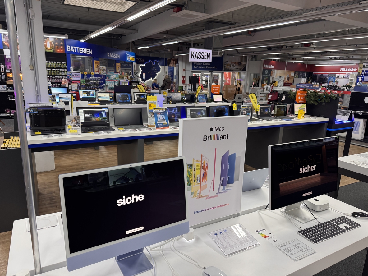 Computer- und Kassenbereich bei EURONICS-Berlet-Soest mit zwei ausgestellten iMacs im Vordergrund, Apple-Werbedisplay in der Mitte und dahinter mehreren Reihen von Laptops sowie Schildern für Batterien und Kassen. Computer- und Kassenbereich bei EURONICS-Berlet-Soest mit zwei ausgestellten iMacs im Vordergrund, Apple-Werbedisplay in der Mitte und dahinter mehreren Reihen von Laptops sowie Schildern für Batterien und Kassen.