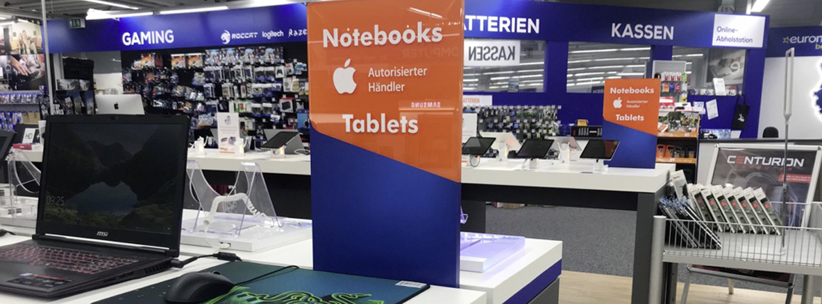 Verkaufsbereich für Notebooks und Tablets bei Euronics Berlat in Soest: Im Vordergrund eine weiße Präsentationstheke mit einem offenen MSI-Gaming-Laptop und einer Gaming-Maus auf einem Mousepad, dahinter zeigt ein hochstehendes, zweifarbiges Schild „Notebooks • Tablets – Autorisierter Apple-Händler“. Im Mittelgrund stehen weitere Tablet-Demogeräte auf weißen Ständern. Im Hintergrund ein blauer „GAMING“-Bereich mit Zubehörwand sowie die blau-weißen Schilder „KASSEN“ und „Online-Abholstation“ über einem Regal mit Batterien und Mobile-Zubehör. Verkaufsbereich für Notebooks und Tablets bei Euronics Berlat in Soest: Im Vordergrund eine weiße Präsentationstheke mit einem offenen MSI-Gaming-Laptop und einer Gaming-Maus auf einem Mousepad, dahinter zeigt ein hochstehendes, zweifarbiges Schild „Notebooks • Tablets – Autorisierter Apple-Händler“. Im Mittelgrund stehen weitere Tablet-Demogeräte auf weißen Ständern. Im Hintergrund ein blauer „GAMING“-Bereich mit Zubehörwand sowie die blau-weißen Schilder „KASSEN“ und „Online-Abholstation“ über einem Regal mit Batterien und Mobile-Zubehör.
