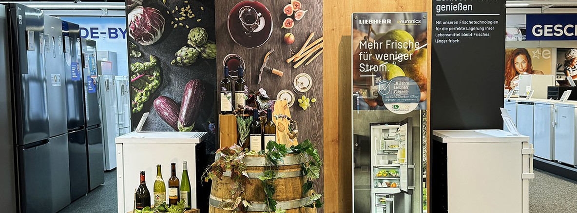 Ausstellungsbereich im EURONICS Berlet Markt mit LIEBHERR-Kühlschrank, Dekoration aus Weinfässern, Weinflaschen und Gemüse; daneben Werbetafel mit dem Slogan „Mehr Frische für weniger Strom“. Ausstellungsbereich im EURONICS Berlet Markt mit LIEBHERR-Kühlschrank, Dekoration aus Weinfässern, Weinflaschen und Gemüse; daneben Werbetafel mit dem Slogan „Mehr Frische für weniger Strom“.
