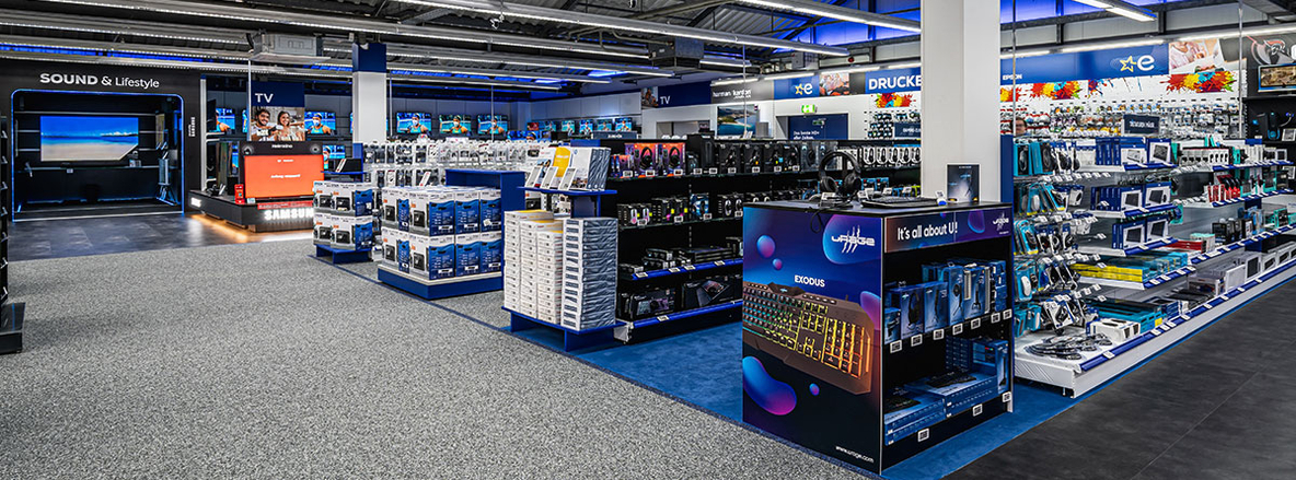 Innenansicht von Euronics-Berlet Bergkamen mit Sound-&-Lifestyle-Demobereich: Im linken Hintergrund eine abgedunkelte Nische mit großem Bildschirm und der Aufschrift „SOUND & Lifestyle“, daneben ein beleuchtetes Samsung-Podest. Im Vordergrund Regale und Präsentationsinseln mit PC-Peripherie, Kopfhörern und Gaming-Zubehör; auf einer Theke prangt ein „Exodus“-Logo mit dem Slogan „It’s all about U!“. Der Boden wechselt von hellem Teppich zu blauem Auslegerteppich und dunkelgrauem Vinyl, die Decke ist offen mit sichtbaren Rohren und Leuchtstofflampen. Innenansicht von Euronics-Berlet Bergkamen mit Sound-&-Lifestyle-Demobereich: Im linken Hintergrund eine abgedunkelte Nische mit großem Bildschirm und der Aufschrift „SOUND & Lifestyle“, daneben ein beleuchtetes Samsung-Podest. Im Vordergrund Regale und Präsentationsinseln mit PC-Peripherie, Kopfhörern und Gaming-Zubehör; auf einer Theke prangt ein „Exodus“-Logo mit dem Slogan „It’s all about U!“. Der Boden wechselt von hellem Teppich zu blauem Auslegerteppich und dunkelgrauem Vinyl, die Decke ist offen mit sichtbaren Rohren und Leuchtstofflampen.