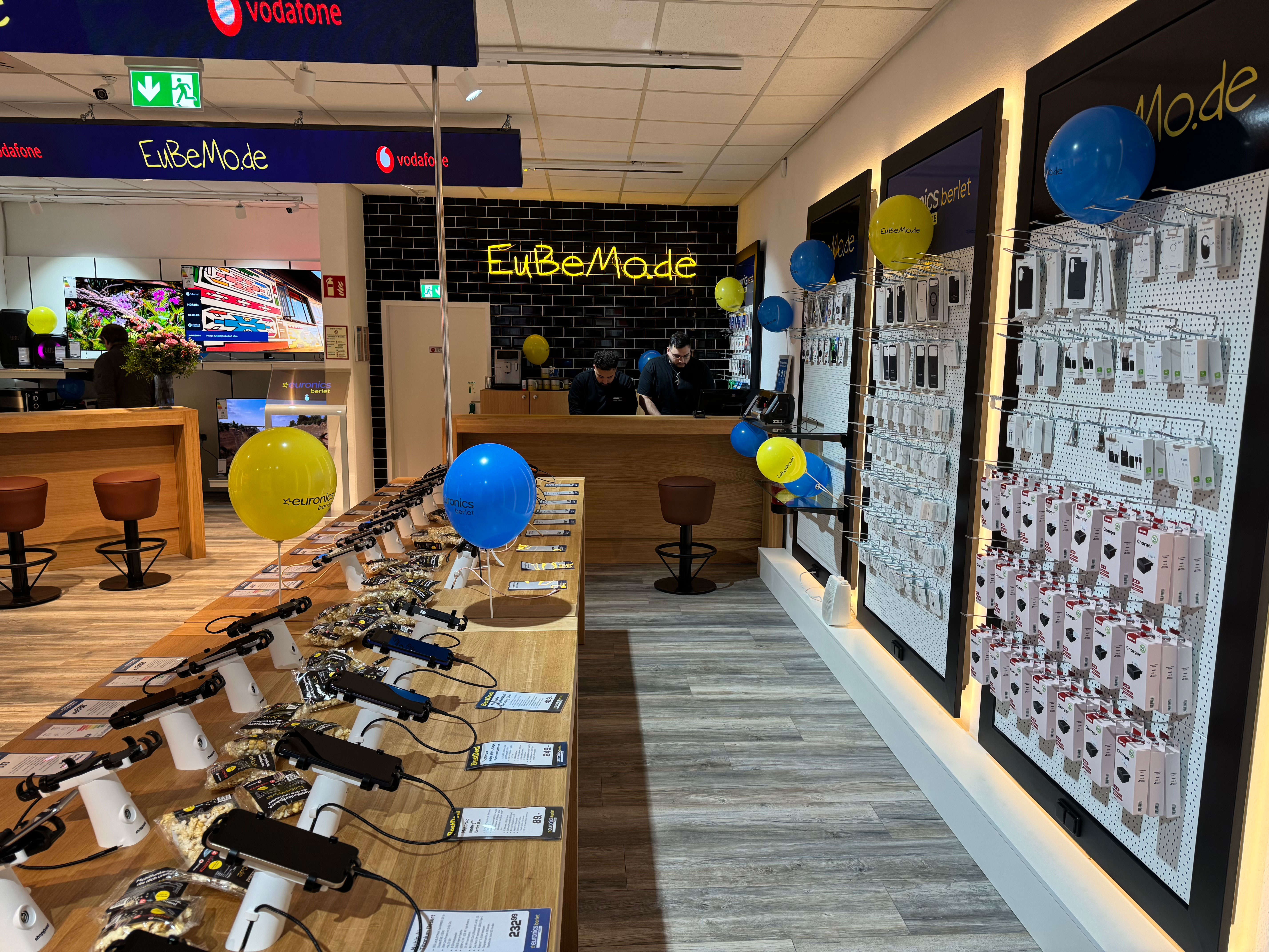 Innenansicht eines modern eingerichteten EURONICS-Berlet-Shops: Holztische mit ausgestellten Smartphones, Zubehörwand rechts, bunte Luftballons und zwei Mitarbeiter an der Theke