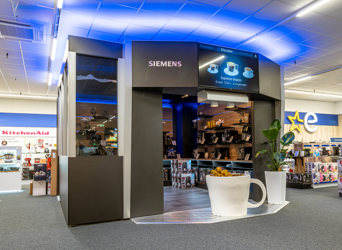 Großer, begehbarer Kaffeevollautomat in der Euronics Berlet Filiale in Hagen mit LED-Beleuchtung. Der Innenraum ist mit Regal ausgebaut auf dem viele verschiedene Kaffeevollautomaten stehen.