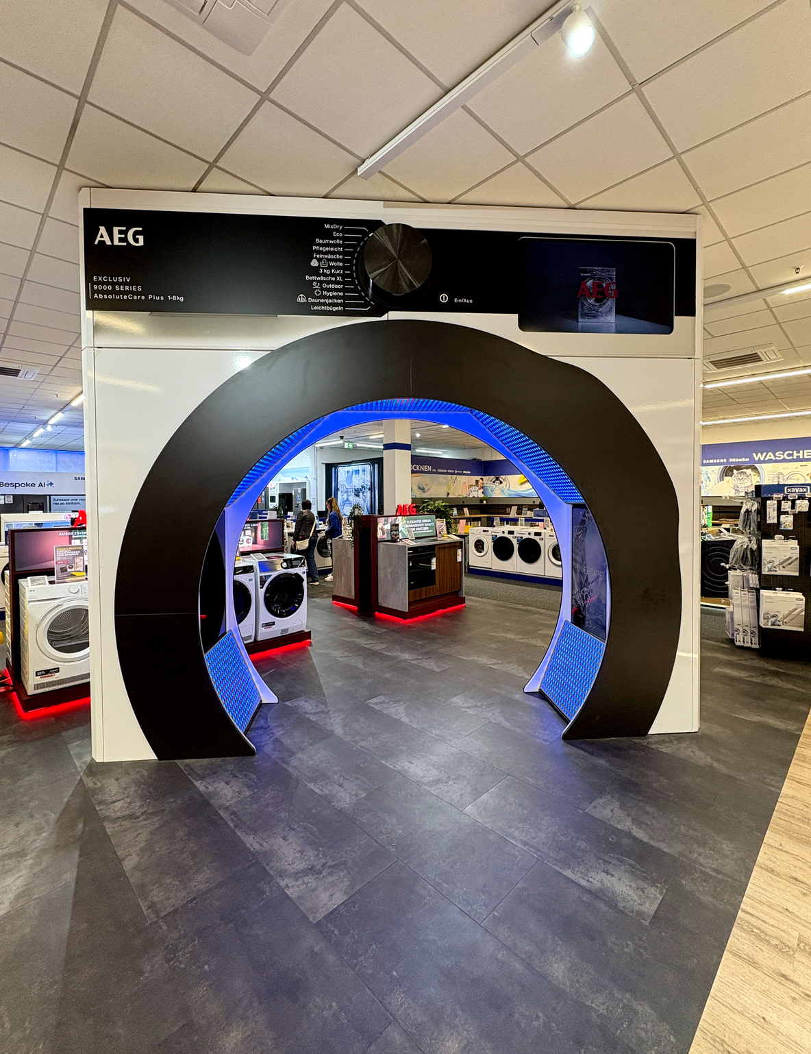 Ausstellung bei Euronics Berlet in Hagen: Eine übermannshohe, begehbare AEG-Installation in Form einer aufgeklappten Waschmaschinentür mit schwarzem Rahmen und blau hinterleuchteter Innenfläche. Oben prangt ein schwarzes Bedienfeld mit AEG-Logo und großem Programmwahlrad. Durch den runden Durchgang sind im Hintergrund Waschmaschinen und Trockner auf dunkelgrauem Boden sowie Theken und Regale unter weißem Raster-Deckensystem zu erkennen.