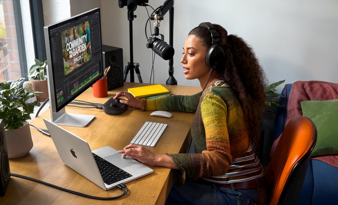 Eine Frau arbeitet an ihrem Schreibtisch zu Hause mit einem MacBook Pro, das mit zwei externen Displays verbunden ist. Sie trägt Kopfhörer und spricht in ein Mikrofon
