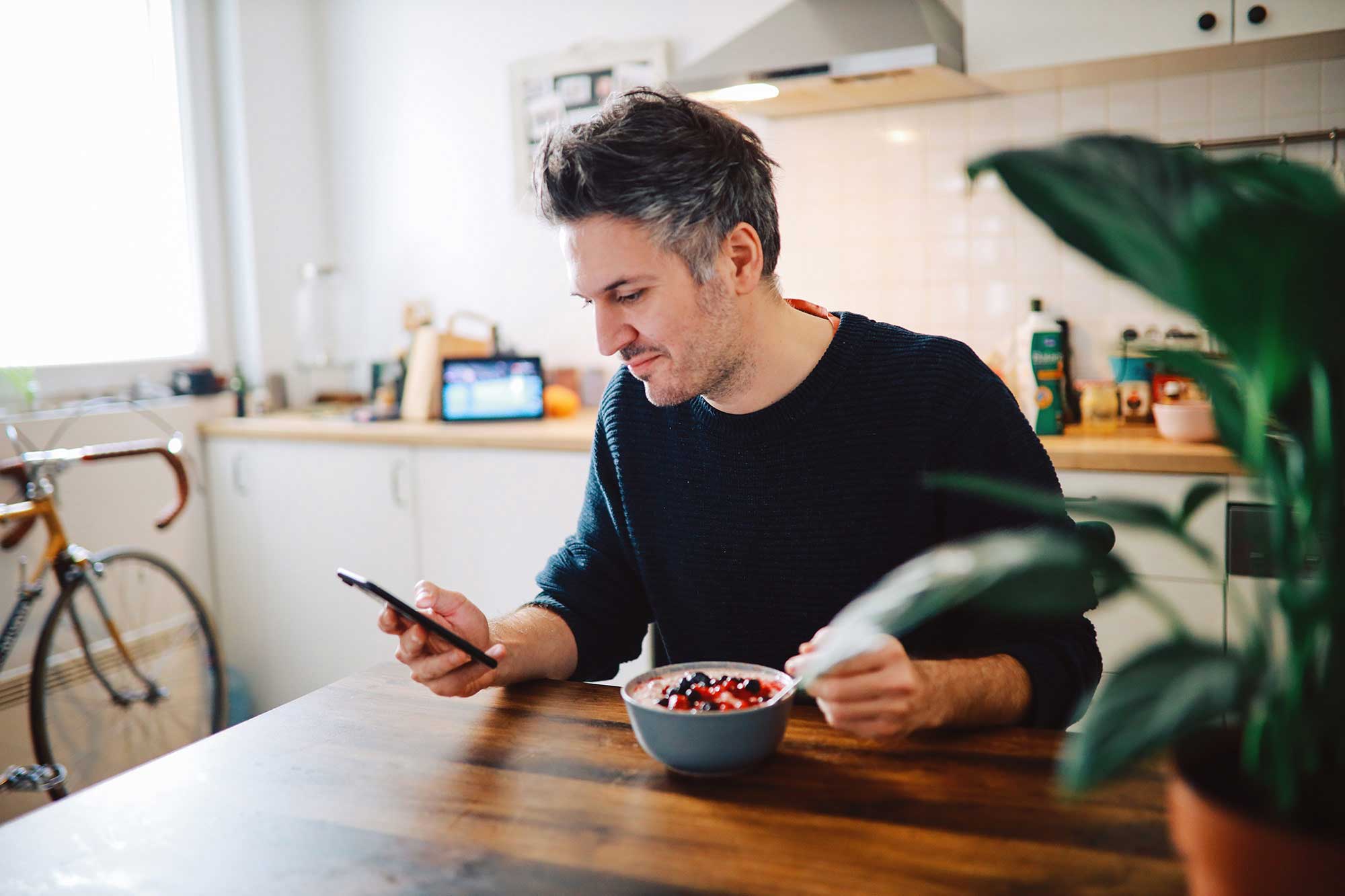 Mann schaut auf Smartphone und frühstückt Müsli